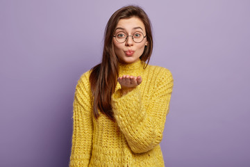 Portrait of lovely dark hared female makes air kiss, sends on distance, expresses love, stretches palm forward, wears glasses winter yellow jumper stands in studio over lilac background. Body language