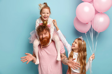 Two funny naughty sisters play together, tickle each other being cared by dad. Cute little girl holds bunch of air balloons. Happy family of father and two daughters prepare for holiday, pose indoor