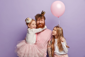 Small girl hugs red haired dad who smiles happily, glad to have two daughters, dressed in festive...