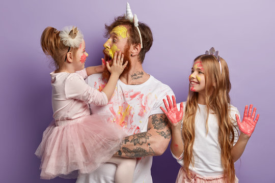 Shocked affectionate dad has dirty face from paints, opens mouth widely, holds little daughter on hands. Smiling girl wears crown, shows palms in pink watercolors. Joyful father and children have fun