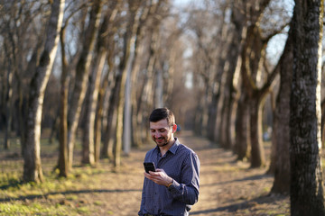 Business bearded man with freckles with short hair in a plaid shirt with watch on his hand looks at the phone and smiles in the autumn forest between the trees in the rays of the sun. Woodland scene.