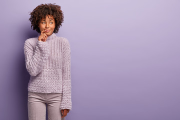 Half length shot of smiling thoughtful Afro Amercan lady looks aside, wears warm winter jumper and trousers in one tone with background, thinks about making shopping on weekened, what to buy
