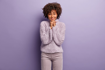 Photo of delighted good looking woman keeps hands under chin, smiles happily, listens attentively pleasant information, wears winter purple sweater, poses indoor. Facial expressions concept.