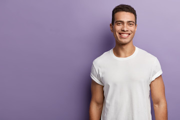 Handsome cheerful young sportsman has sporty body, muscular arms, wears white mock up t shirt, has short dark hair, toothy appealing smile, stands over purple background, blank copy space aside