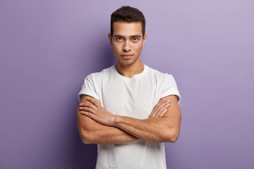 Self assertive serious young guy keeps muscular arms crossed over chest, looks directly at camera, gazes attentively something, dressed in casual white t shirt, looks confident. I am ready listen you