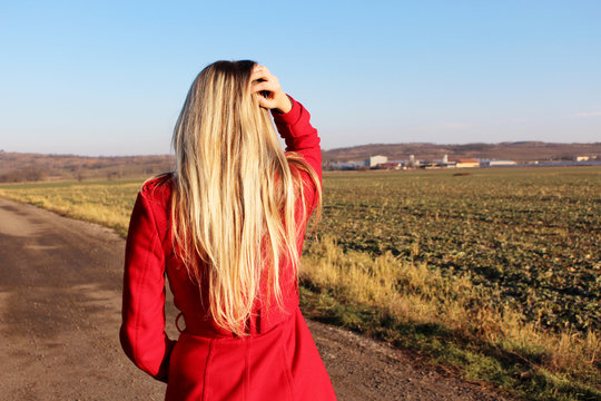 Young beautiful blonde girl in red coat standing on a dirt road thinking about another way. Traveler background.