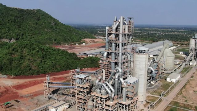 Aerial drone view orbiting industrial tower and cement processing plant in Asia, with massive ducts, pipes, and storage sheds visible at foot of low mountains.