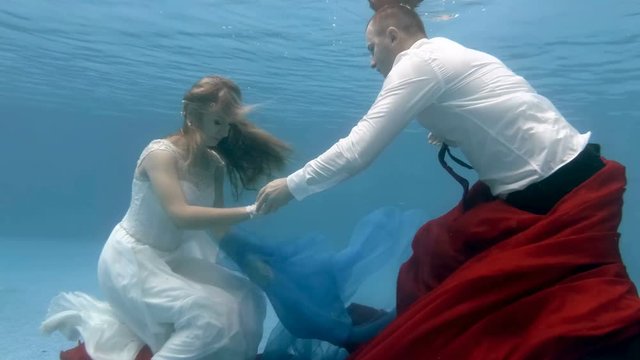 The bride in white dress and the groom swim and play underwater in the pool with red and blue fabric. Wedding underwater. Portrait. Zoom. 4K. 29.97 fps