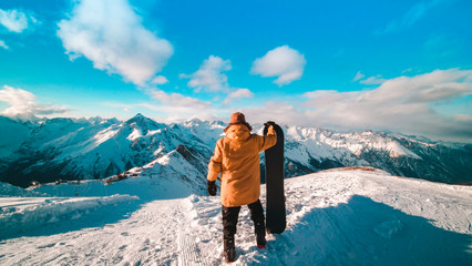 A young guy in a yellow down jacket with a snowboard in his hands stands on the top of the...