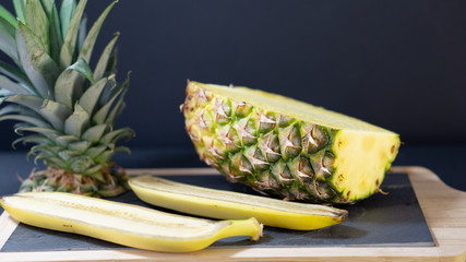 A pineapple half on a stone-framed stone board is cut in half, about two halves of a banana, the pineapple tip is sliced next to it, the background is black