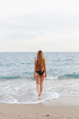 Young slim beautiful tanned woman with long blonde hair in black swimsuit walking on the beach. View from the back.