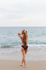 Fototapeta premium Młoda szczupła piękna opalona kobieta z długimi blond włosami w czarnym stroju kąpielowym spaceru na plaży. Widok z tyłu.
