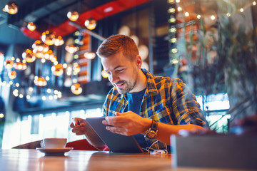 Smiling handsome Caucasian unshaven man in plaid shirt using tablet in cafeteria. In front of him on desk coffee.