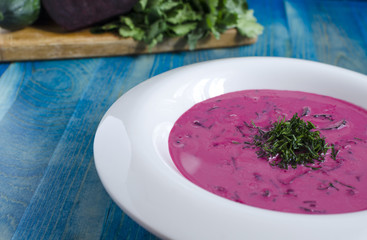 Cold soup of beetroot and vegetables with herbs on a white plate on blue background of field foreground