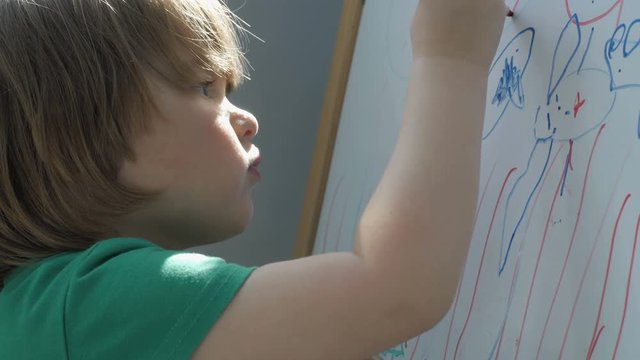 Child painting on easel. Closeup. Cute little boy is drawing with pencils in preschool. Child draw with colorful markers and smile.