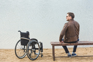 A young man sits on a bench by the lake, next to his wheelchair.