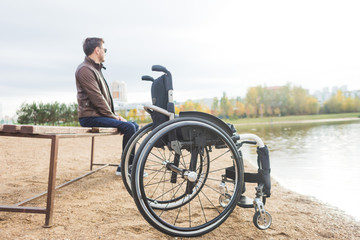 A young man sits on a bench by the lake, next to his wheelchair.