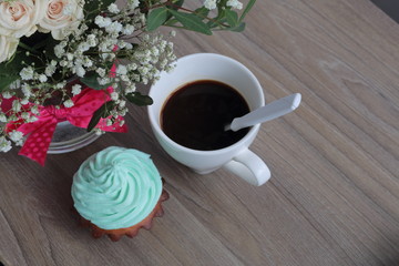 Cream cake and cup of coffee. A coffee spoon is dropped into the coffee cup. Nearby is a bouquet of flowers in a glass vase.
