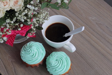 Two cream cakes and cup of coffee. A coffee spoon is dropped into the coffee cup. Nearby is a bouquet of flowers in a glass vase.