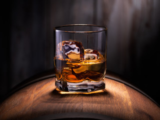 Glass of whiskey with ice cubes on the wooden barrel with wooden background - obrazy, fototapety, plakaty