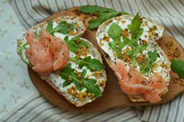 Homemade salmon sandwiches with cream cheese, mustard seeds, olive oil, arugula on toasted rye bread, served on the wooden board. Healthy trendy food. Table napkin with grey lines.