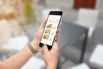 Female hands ordering food with food delivery app on black smartphone, blurred dining room in background - obrazy, fototapety, plakaty