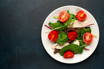 Mix fresh leaves of arugula, lettuce, spinach, tomato and chicken fillet for salad, on a white plate on a stone background with copy space for your text