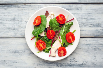 Mix fresh leaves of arugula, lettuce, spinach, tomato and chicken fillet for salad, on a white plate on a wood background
