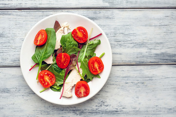 Mix fresh leaves of arugula, lettuce, spinach, tomato and chicken fillet for salad, on a white plate on a wood background