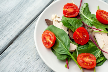 Mix fresh leaves of arugula, lettuce, spinach, tomato and chicken fillet for salad, on a white plate on a wood background