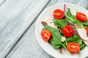 Mix fresh leaves of arugula, lettuce, spinach, tomato and chicken fillet for salad, on a white plate on a wood background