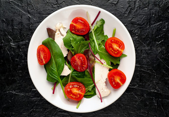 Mix fresh leaves of arugula, lettuce, spinach, tomato and chicken fillet for salad, on a white plate against a stone