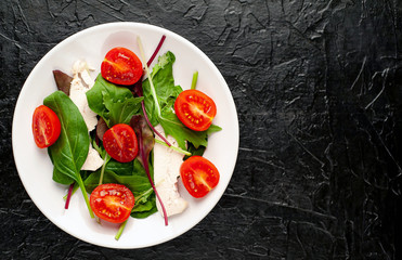 Mix fresh leaves of arugula, lettuce, spinach, tomato and chicken fillet for salad, on a white plate against a stone