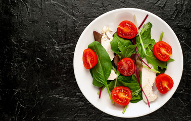 Mix fresh leaves of arugula, lettuce, spinach, tomato and chicken fillet for salad, on a white plate against a stone