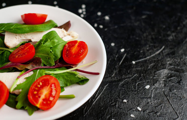 Mix fresh leaves of arugula, lettuce, spinach, tomato and chicken fillet for salad, on a white plate against a stone