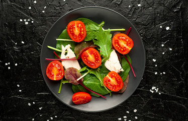 Mix fresh leaves of arugula, lettuce, spinach, tomatoes and chicken fillet for salad, on a dark plate against a background of concrete
