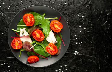 Mix fresh leaves of arugula, lettuce, spinach, tomatoes and chicken fillet for salad, on a dark plate against a background of concrete
