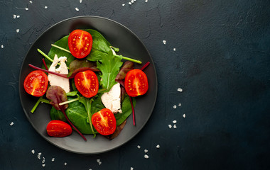 Mix fresh leaves of arugula, lettuce, spinach, tomatoes and chicken fillet for salad, on a dark plate against a background of concrete