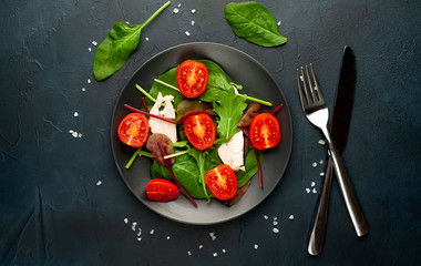 Mix fresh leaves of arugula, lettuce, spinach, tomatoes and chicken fillet for salad, on a dark plate against a background of concrete