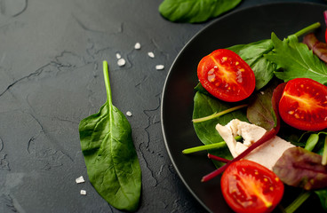 Mix fresh leaves of arugula, lettuce, spinach, tomatoes and chicken fillet for salad, on a dark plate against a background of concrete