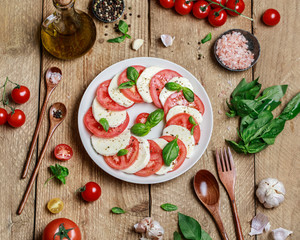 Delicious italian caprese salad with ripe tomatoes, fresh basil and mozzarella cheese on wooden rustic background. Italian caprese salad in white plate with sliced tomatoes.