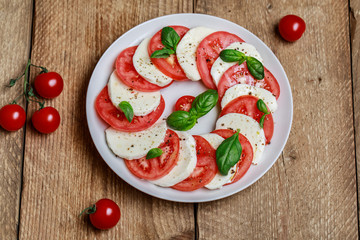 Delicious italian caprese salad with ripe tomatoes, fresh basil and mozzarella cheese on wooden rustic background. Italian caprese salad in white plate with sliced tomatoes.