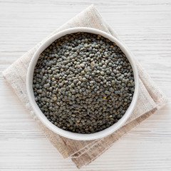 Dry green french lentils in a bowl over white wooden background. Flat lay, top view, from above. Close-up.