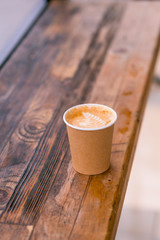 A paper coffee cup with cappuccino in the street cafe on the wooden background. Place for text.