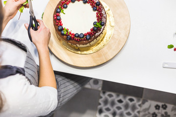 Confectioner decorates with berries a biscuit cake with white cream and chocolate. Cake stands on a wooden stand on a white table. The concept of homemade pastry, cooking cakes.