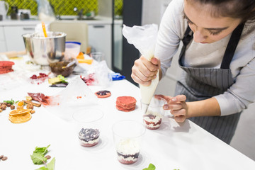 A confectioner prepares a trifle in three cups. Desserts are on the white table in the kitchen. The concept of homemade pastry, cooking cakes.
