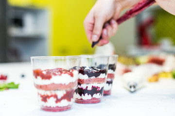 A confectioner prepares a trifle in three cups. Desserts are on the white table in the kitchen. The concept of homemade pastry, cooking cakes.