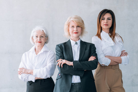 Successful business women. Ambitious female team. Confident mature and young ladies standing with arms folded over light wall.