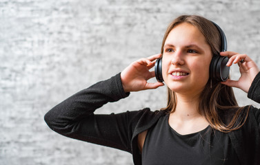 portrait of young teenager brunette girl with long hair listening music on wireless headphones on gray wall background