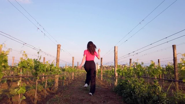 Bella ragazza con maglia rosa e pantaloni neri corre felice in mezzo ai vigneti in campagna.